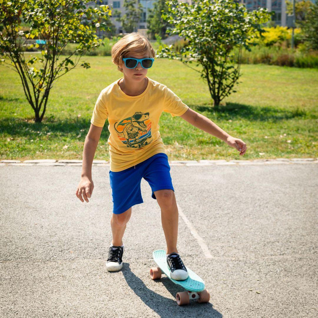 young boy wearing yellow tee with a dinosaur on it with a pair of bright blue shorts while on a skateboard. 