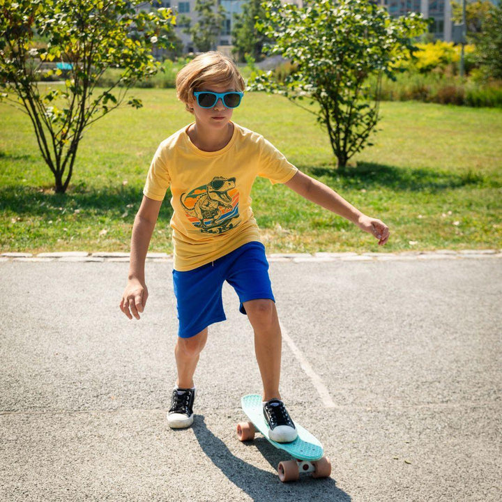 young boy wearing yellow tee with a dinosaur on it with a pair of bright blue shorts while on a skateboard. 