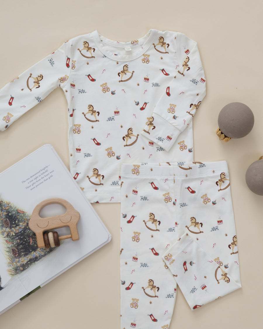 A baby long-sleeve pajama set with matching pants, featuring an all-over playful print of rocking horses, teddy bears, high heels, and small red bows on a warm cream background. The set is styled in a flat-lay arrangement with a wooden toy, an open book, and two gray ornaments on a light beige background.