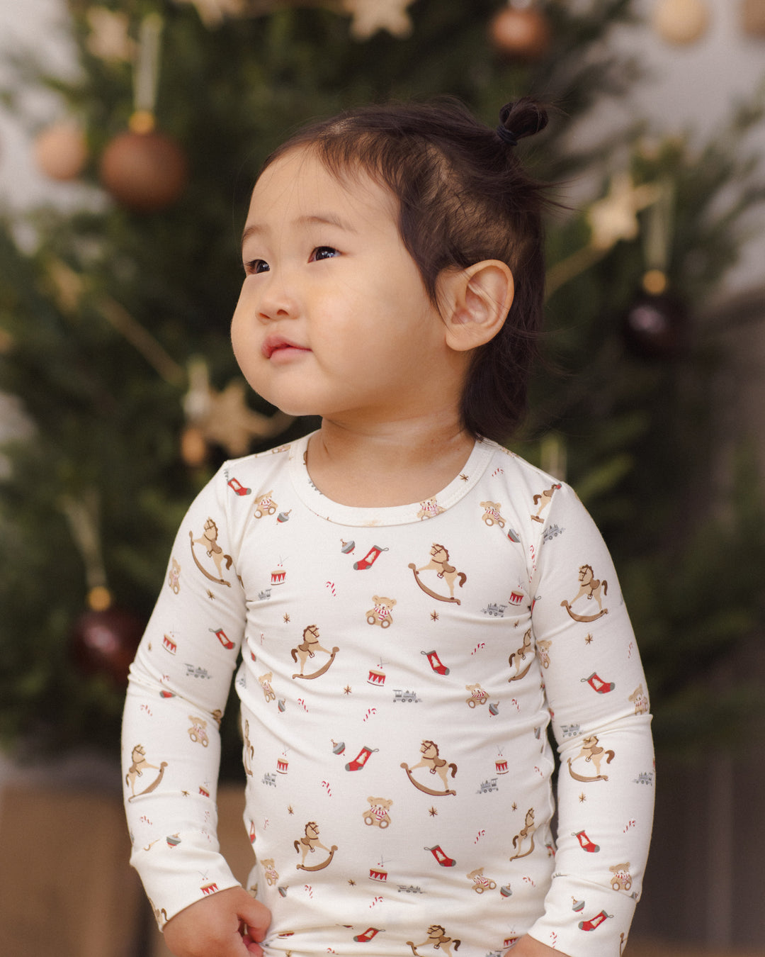 A toddler girl wearing a white long-sleeve pajama top with a Christmas-themed print of rocking horses, candy canes, and ornaments, standing in front of a blurred Christmas tree.