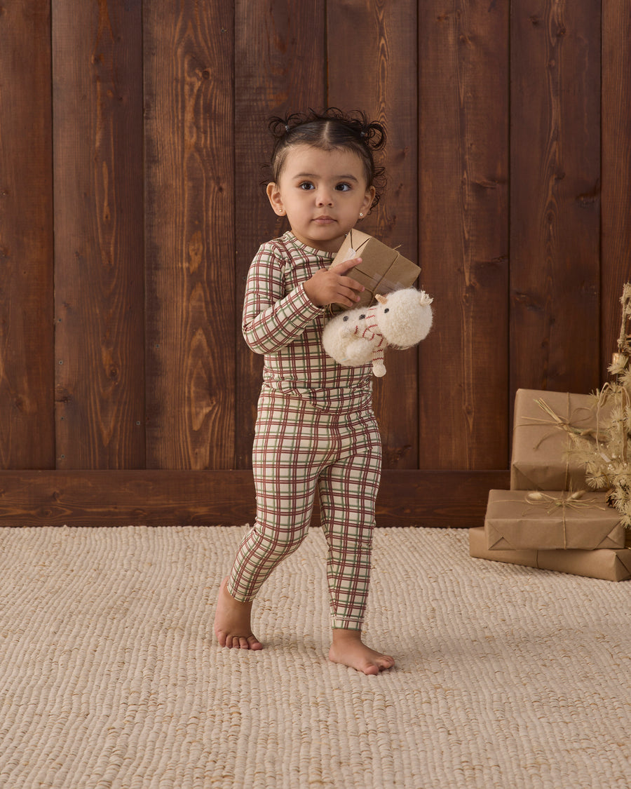 A toddler stands barefoot on a textured beige rug in front of a dark wood plank wall, wearing a brown and beige plaid long-sleeve pajama set and holding a small brown gift box with a white plush toy.