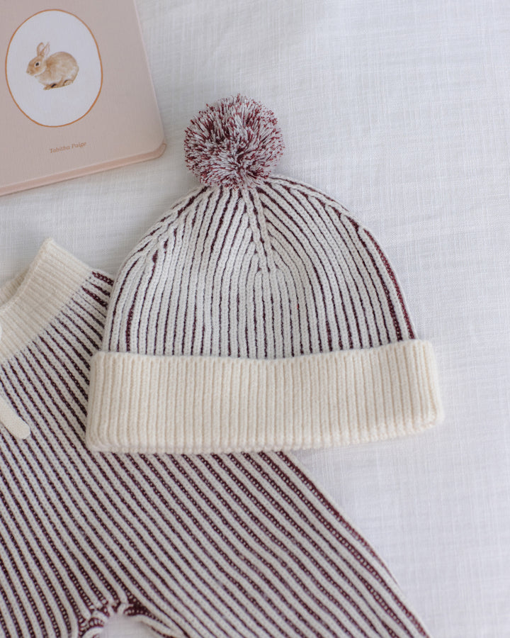 A ribbed knit baby beanie with a pom-pom, featuring alternating cream and dark brown stripes, styled on a white textured fabric with a matching striped garment and a book in the background.
