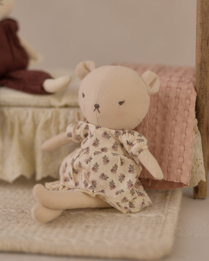 A small beige plush teddy bear doll wearing a cream dress with a muted floral print, posed on a woven rug in front of a pink quilted blanket.