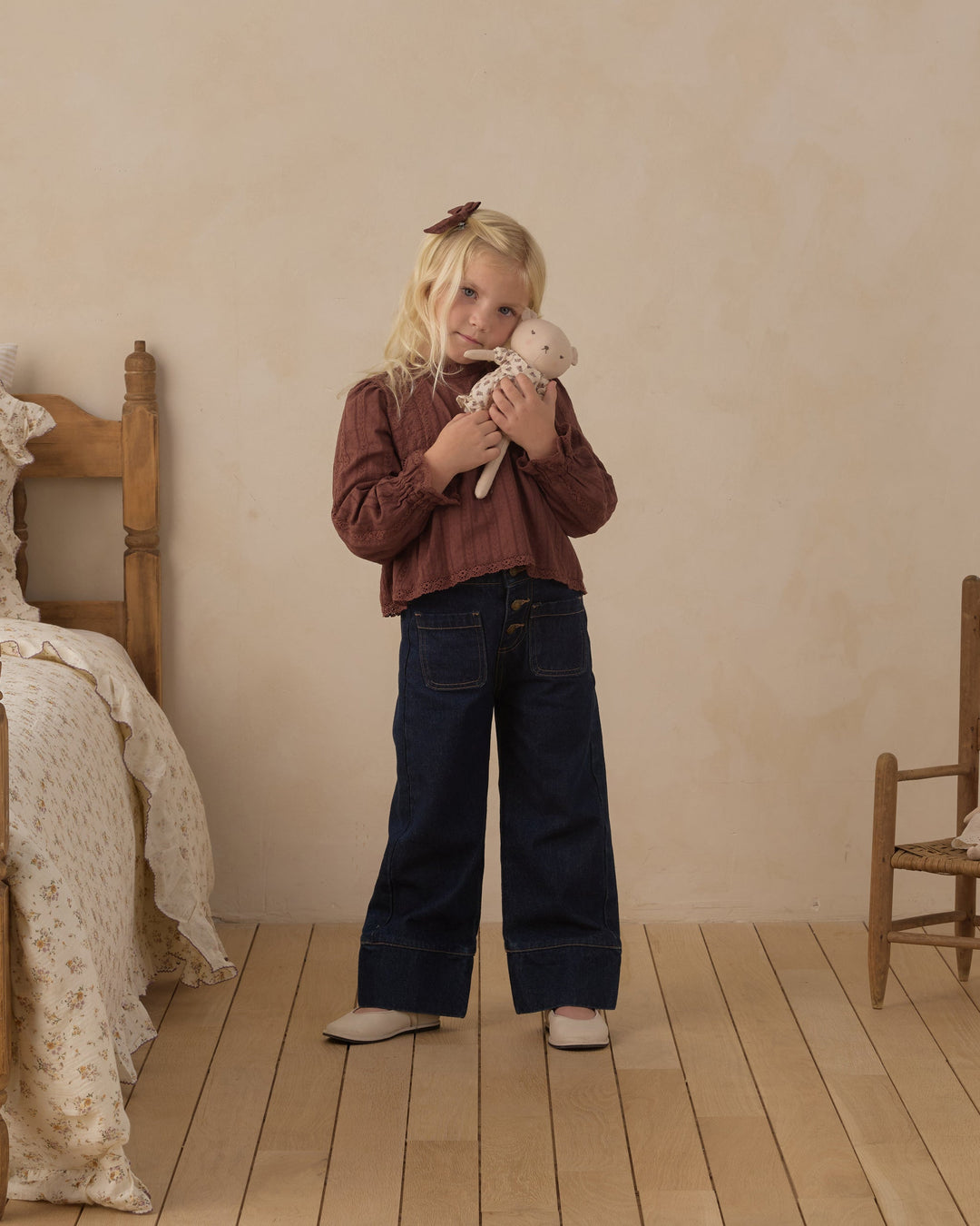 A young blonde girl stands in a rustic bedroom setting, wearing a brown ribbed long-sleeve top and dark wide-leg jeans, holding a small white stuffed animal.