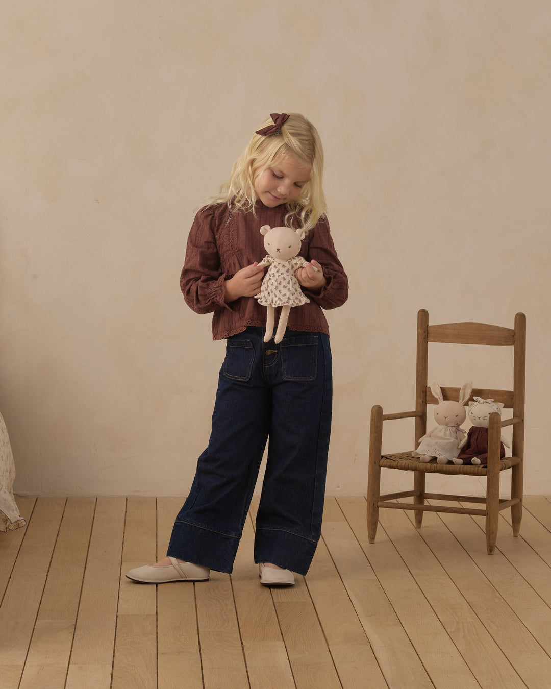 A young girl stands in a room with a beige wall and light wood floor, wearing a brown long-sleeve top and dark blue wide-leg pants while holding a small cream teddy bear. A wooden chair with stuffed toys sits to her right.