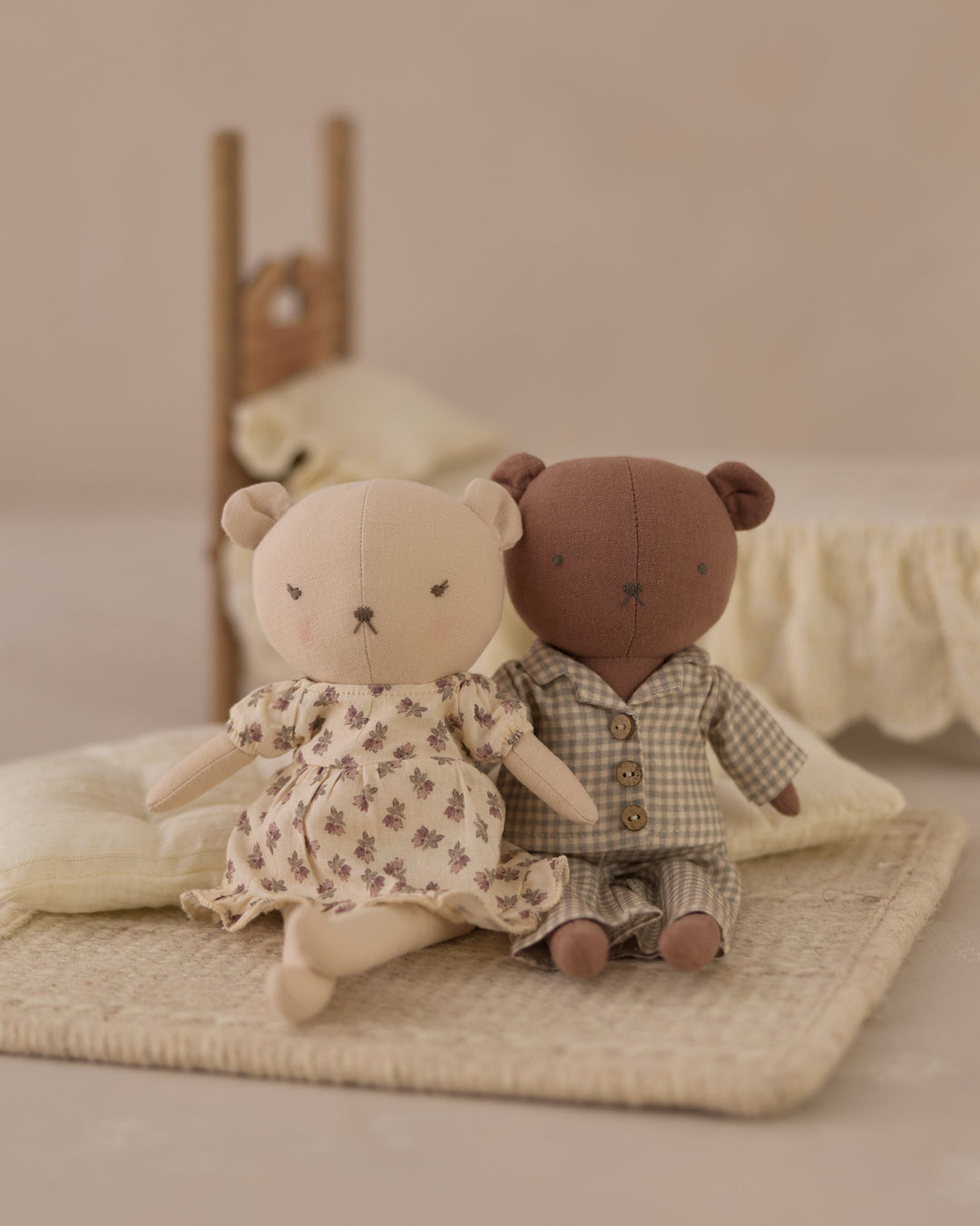 Two small teddy bear plush toys sitting together on a woven mat. One bear is cream-colored wearing a cream floral dress, and the other is brown wearing a brown-and-white checkered pajama set.