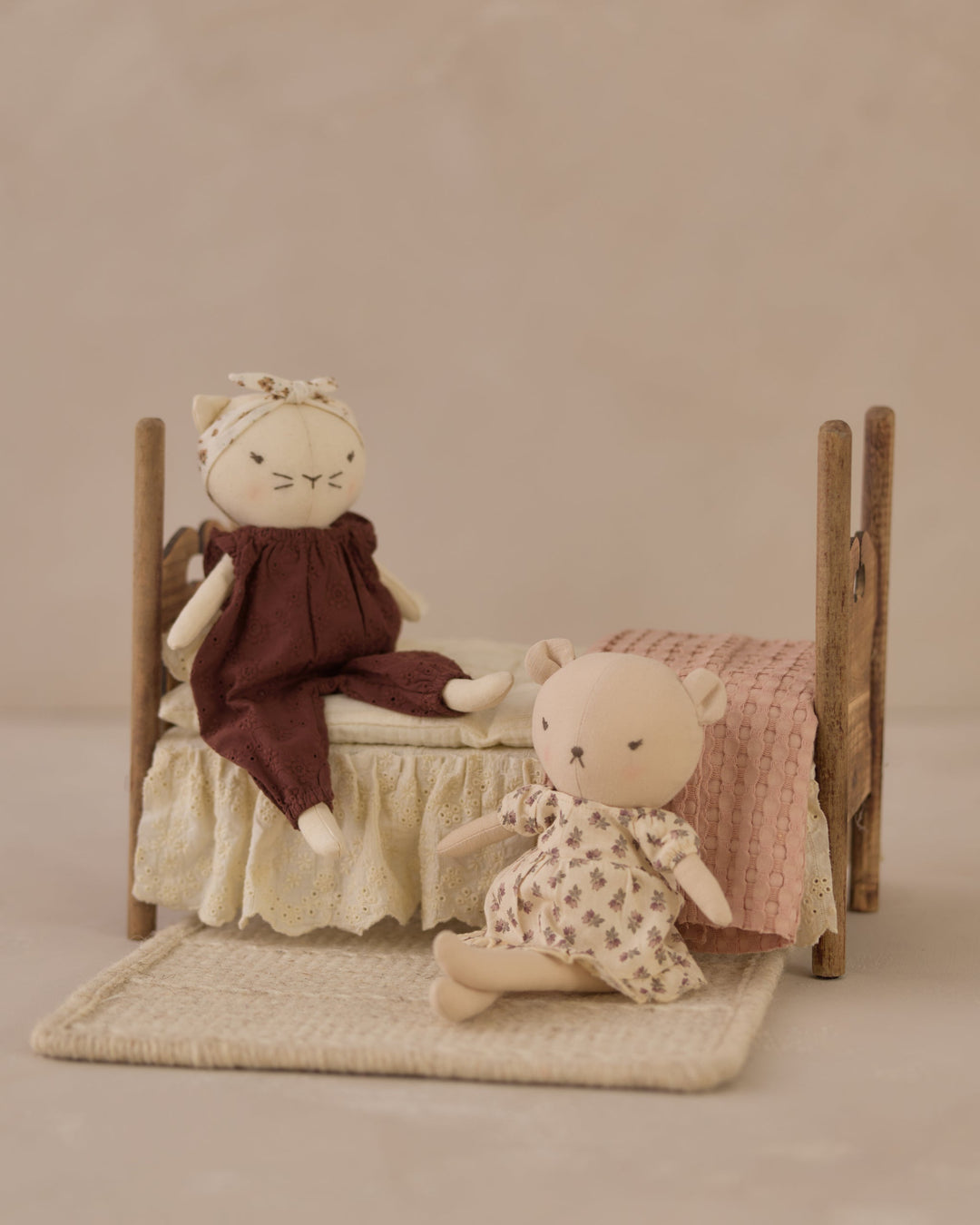 A miniature dollhouse-style bed with a cream ruffled mattress and bedding, a small plush doll in a dark brown dress sitting on the bed, and another small plush doll in a cream patterned outfit beside it on a woven rug against a soft beige background.