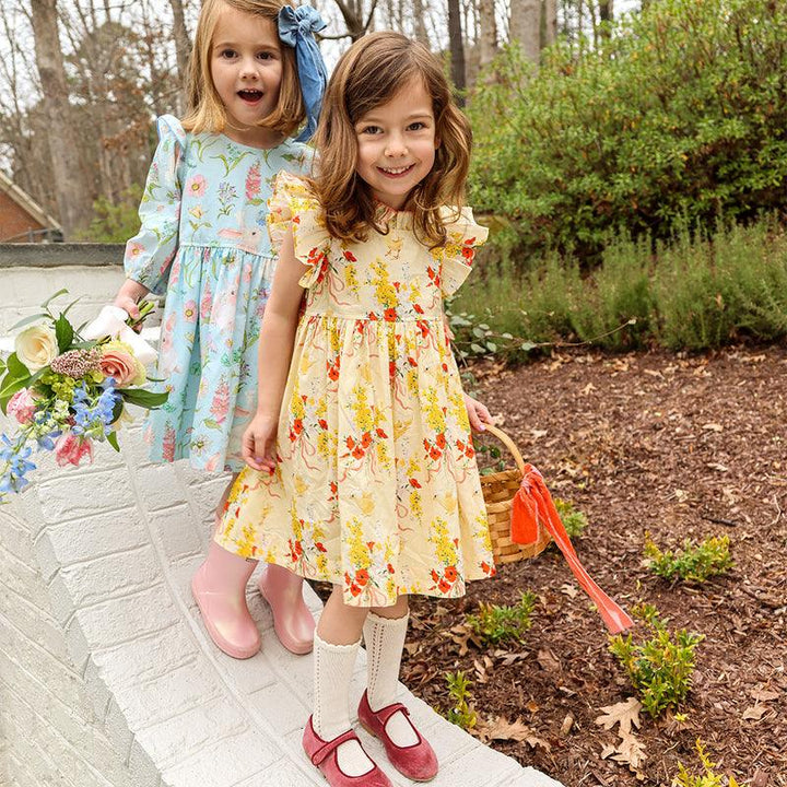 two little girls wearing Easter dresses, one yellow with chicks and flowers on it  and the other pale blue with bunnies and light pink flowers. 