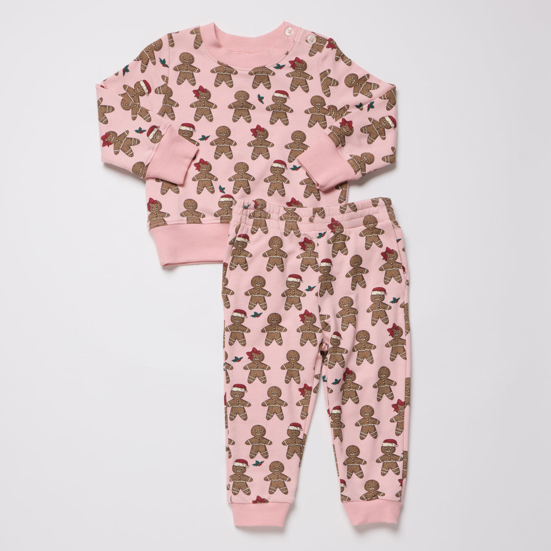 A two-piece children's pajama set featuring a long-sleeve top and matching jogger pants in light pink, both covered in a repeating print of gingerbread cookies wearing small holiday hats and scarves.
