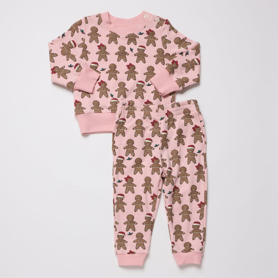 A two-piece children's pajama set featuring a long-sleeve top and matching jogger pants in light pink, both covered in a repeating print of gingerbread cookies wearing small holiday hats and scarves.