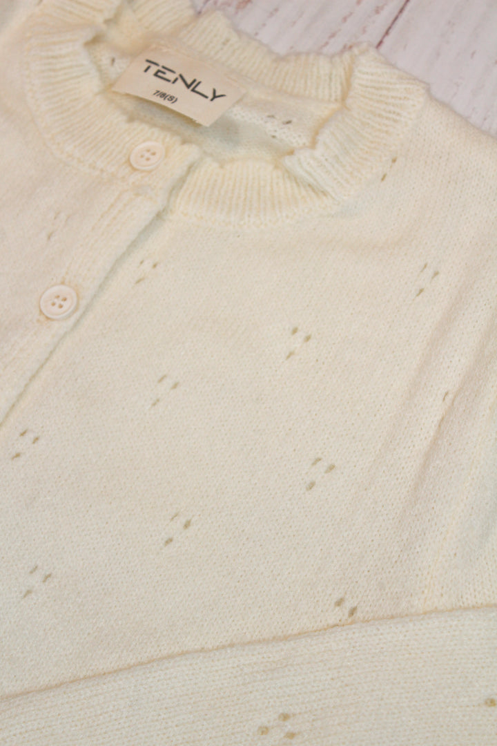 Close-up of a cream-colored cardigan with a brand label on a light wooden surface.