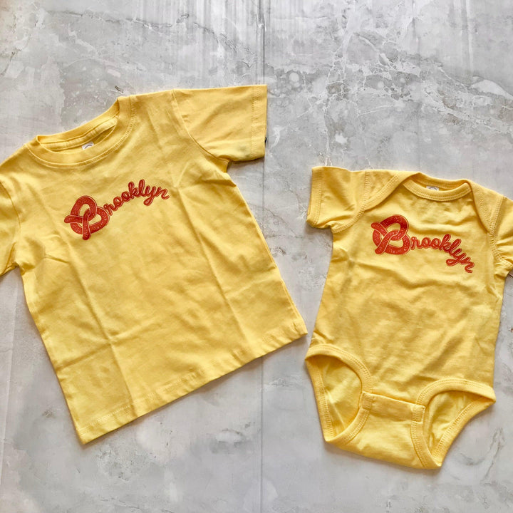 matching baby and kid tee and onesies with Brooklyn written on it. a pretzel being the "B" on yellow tee or onesie