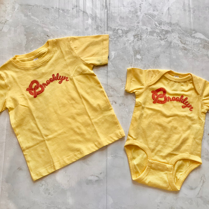 matching baby and kid tee and onesies with Brooklyn written on it. a pretzel being the "B" on yellow tee or onesie