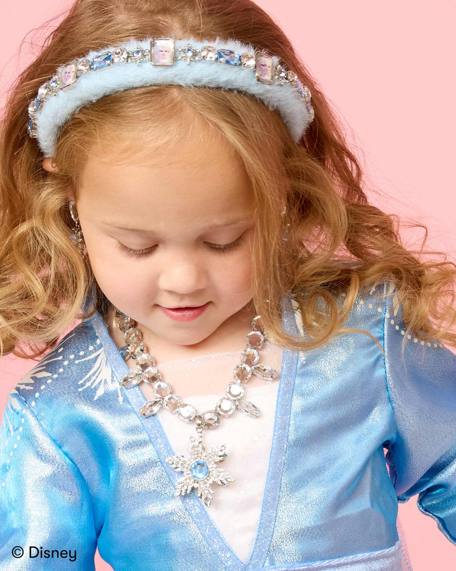 Child in a blue costume with a Frozen headband and necklace on a pink background