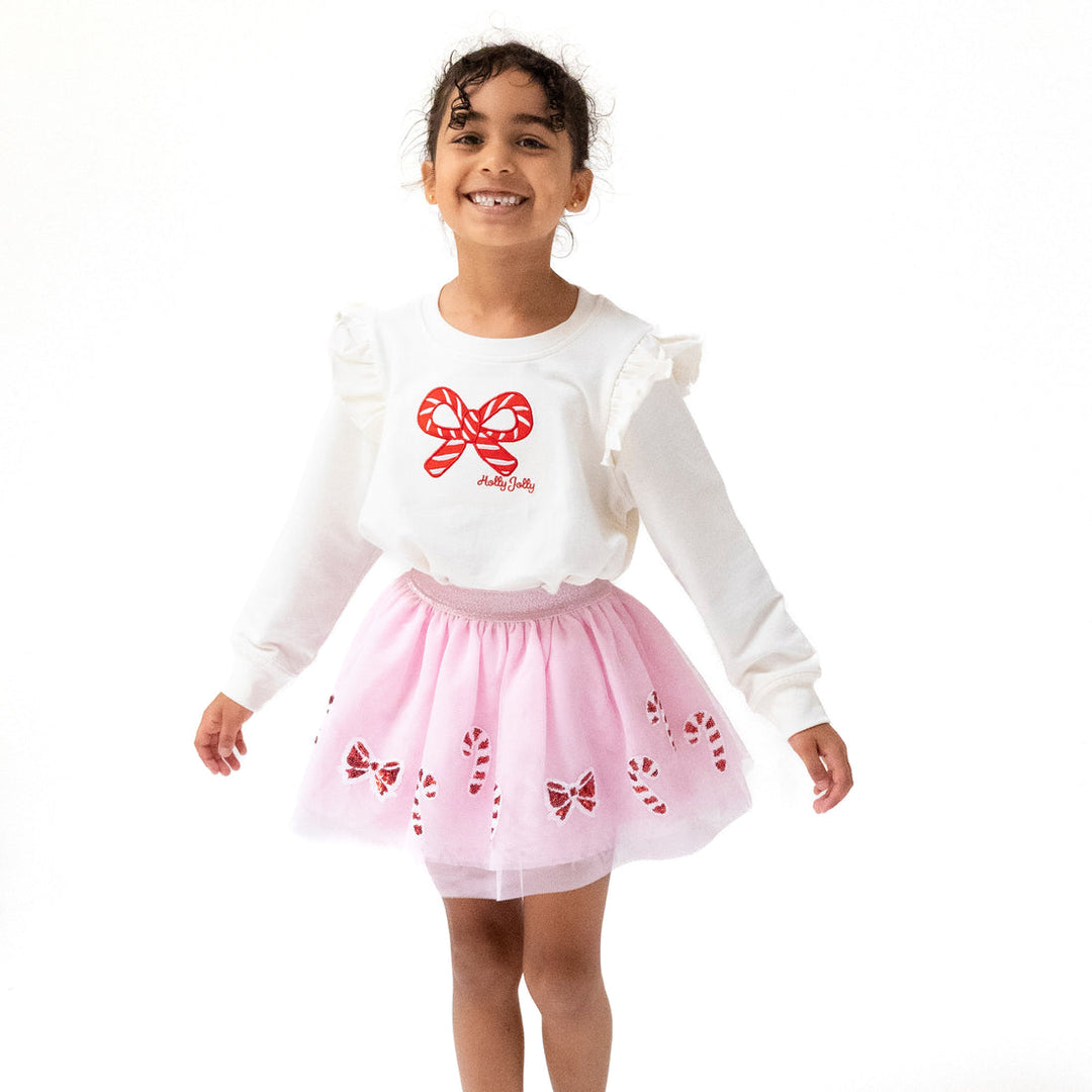 Young girl wearing a white sweater with a red candy cane design and a pink skirt with bow patterns on a white background.