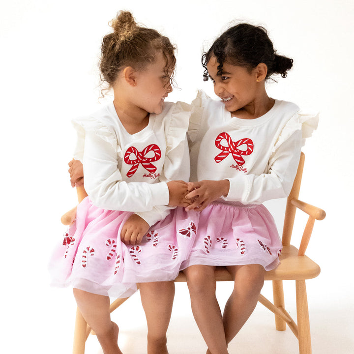 Two young girls wearing matching outfits with red bows and pink skirts sitting on a chair.