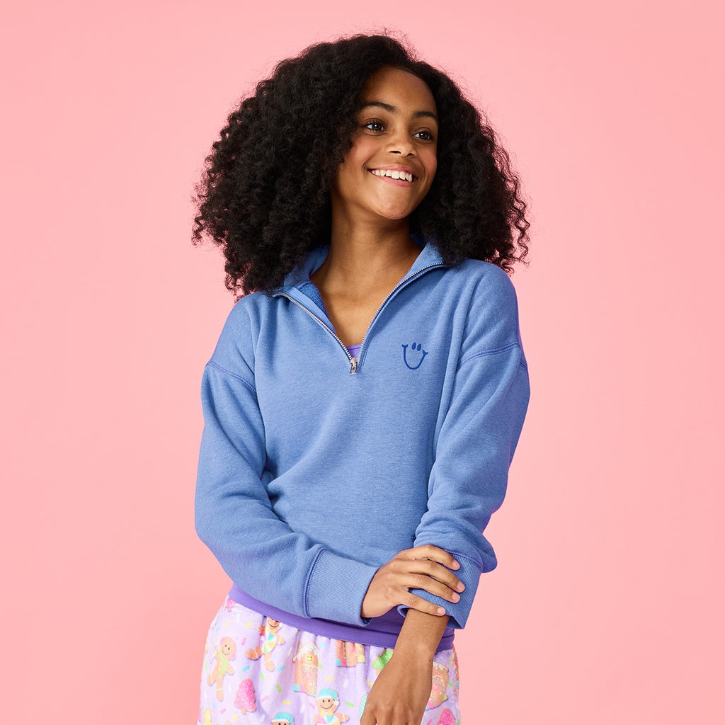 A woman with curly black hair wearing a heathered blue quarter-zip sweatshirt and pastel patterned pajama pants, posing against a solid pink background.
