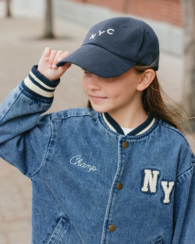 Child wearing a navy cap with 'NYC' and a denim jacket with 'Champ' and 'NY' patches.