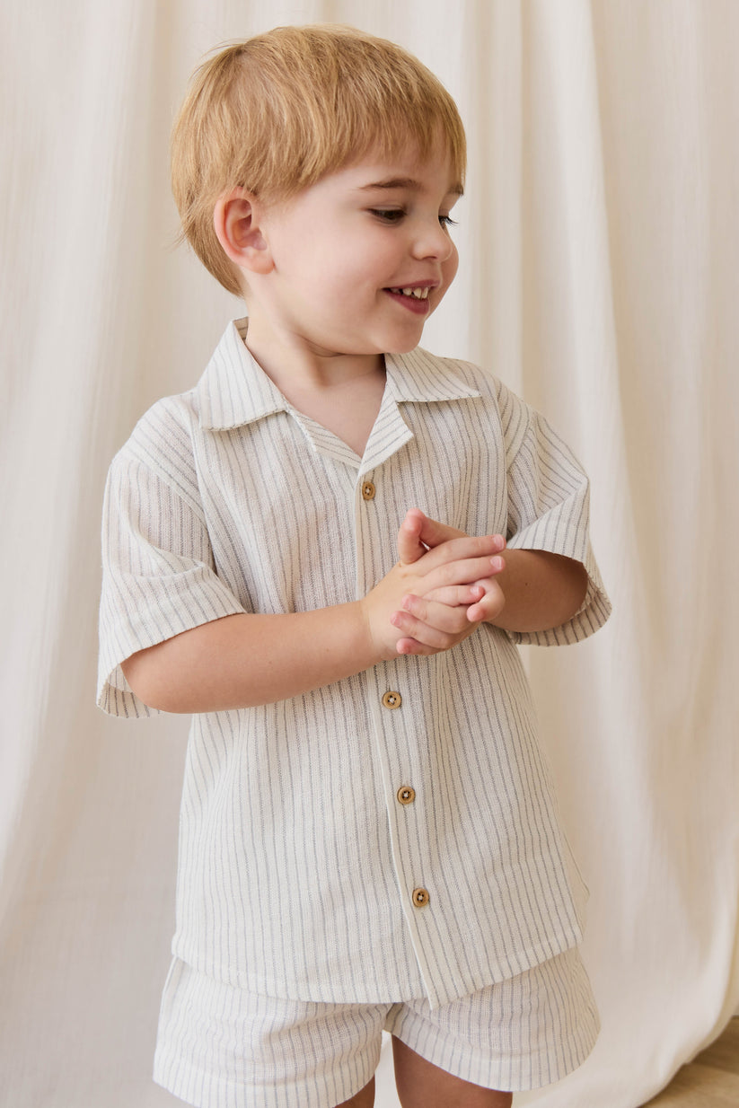 Child wearing a light-colored button-up shirt and shorts against a white curtain background