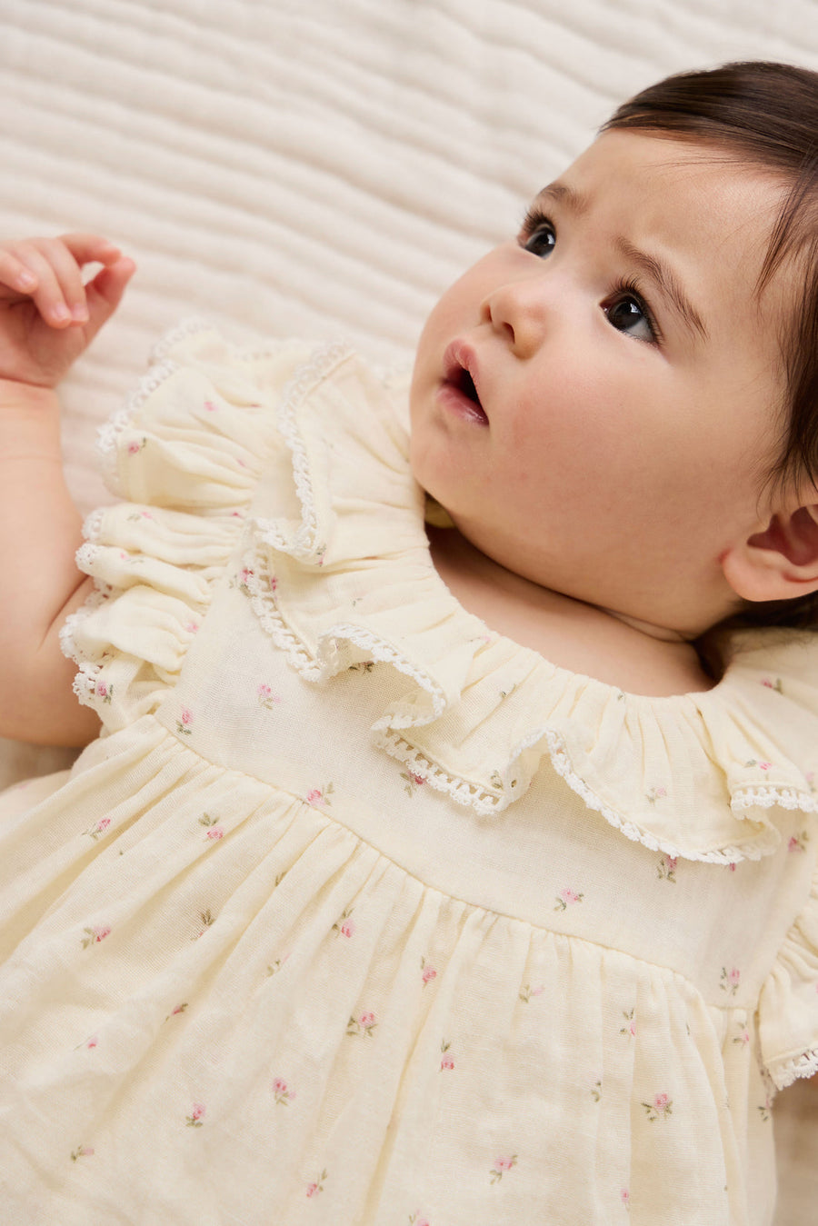 Baby in a cream-colored dress with floral patterns lying on a textured surface.