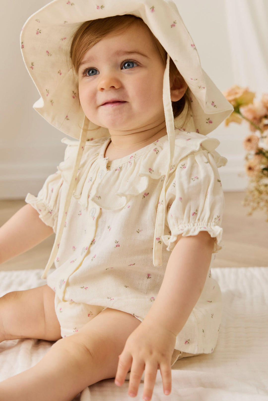 Baby wearing a floral hat and outfit sitting on a soft surface.