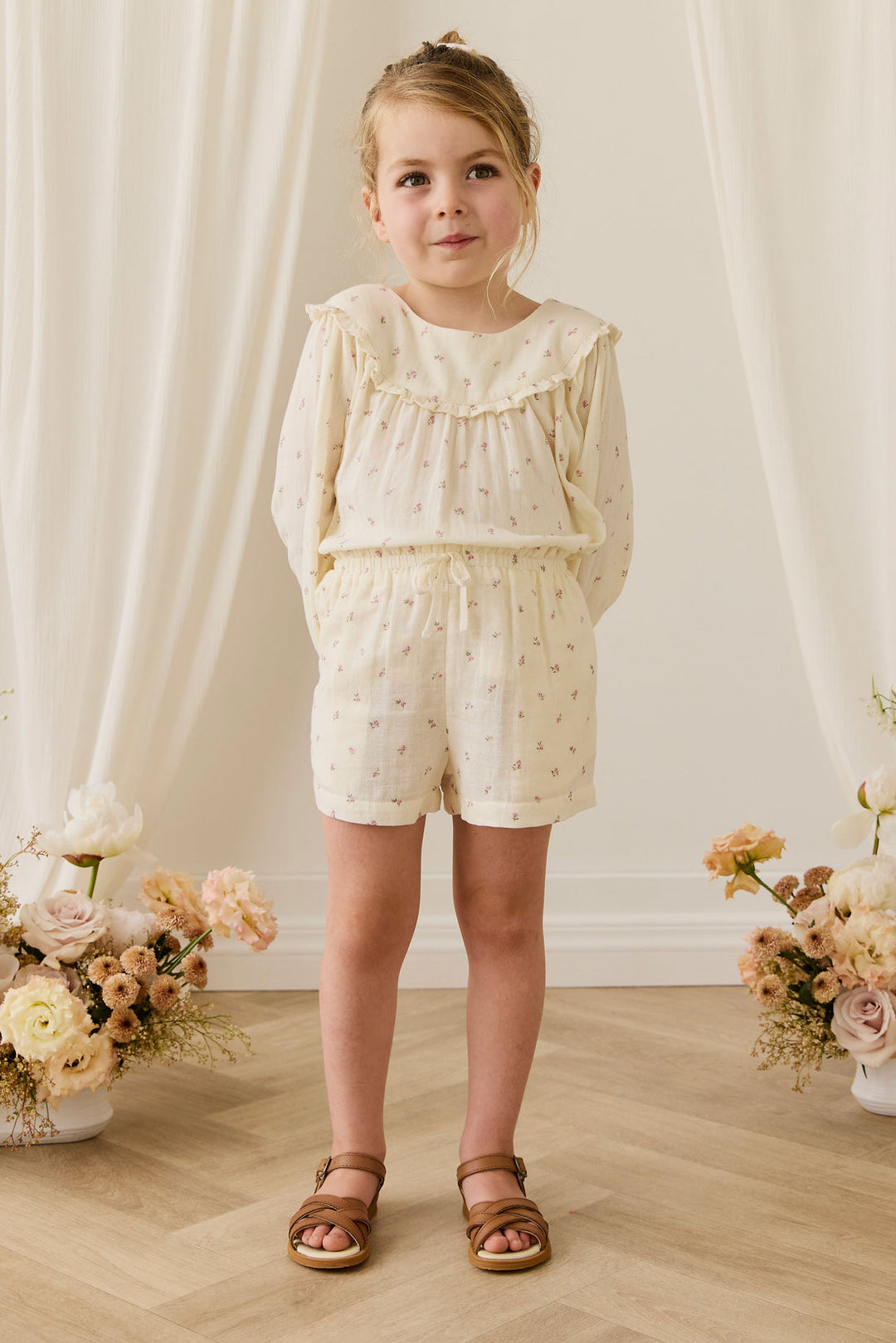Young girl in a white outfit standing in front of a white curtain with floral arrangements on either side.