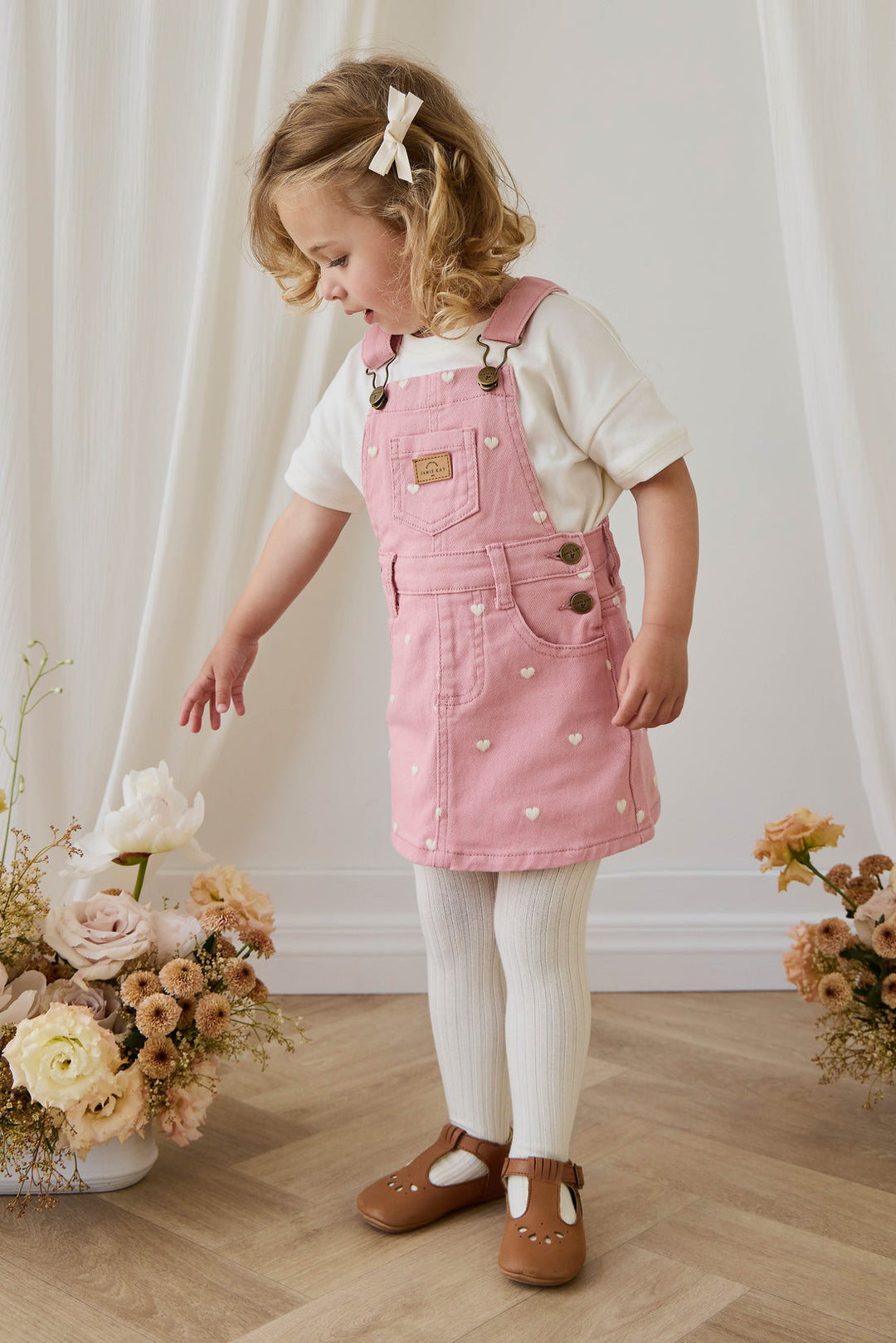 Young girl wearing a pink pinafore dress with white polka dots, standing in a room with floral arrangements.