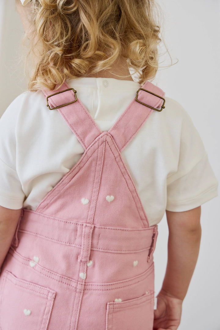 Person wearing pink overalls with a white shirt underneath on a plain background
