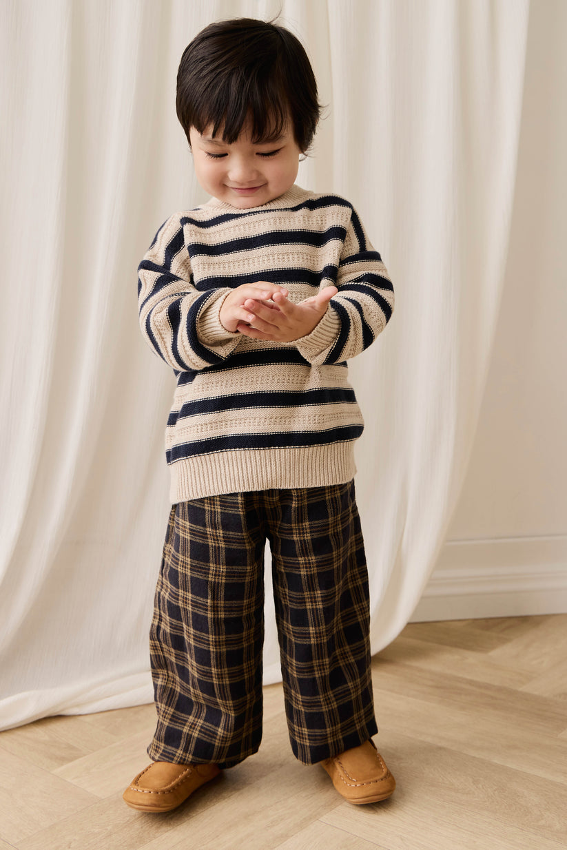 Child wearing a striped sweater and plaid pants standing against a white curtain.