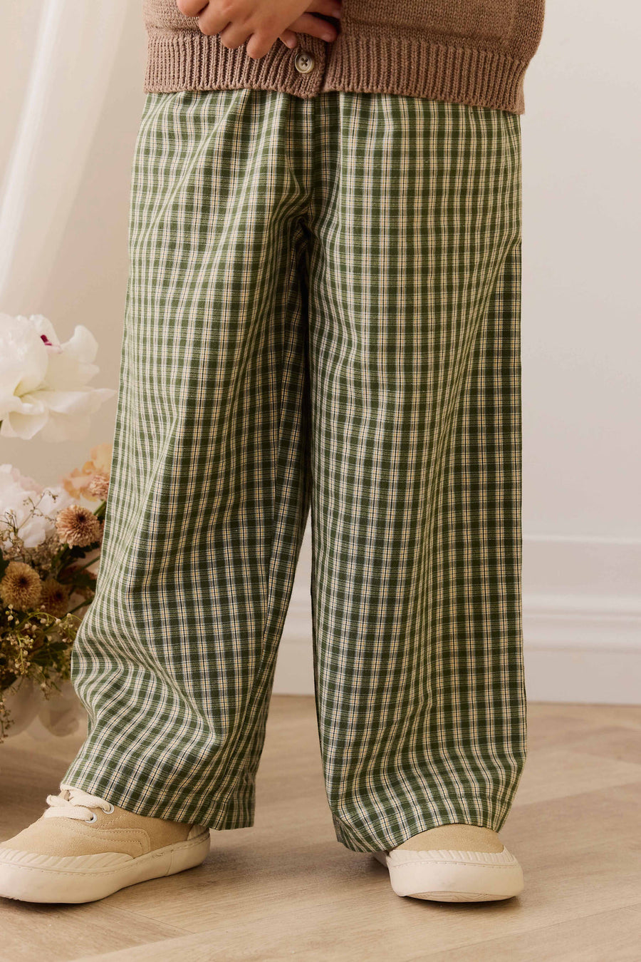 kid wearing green checkered pants and a brown sweater in a room with flowers in the background.