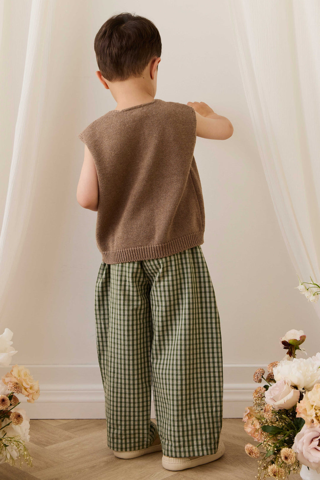 Child wearing a brown vest and green checkered pants standing in a softly lit room with floral decorations.