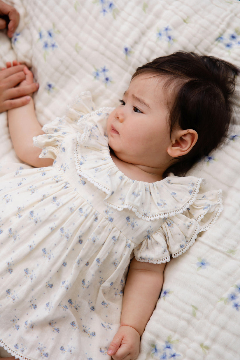Baby in a white floral dress lying on a quilted surface