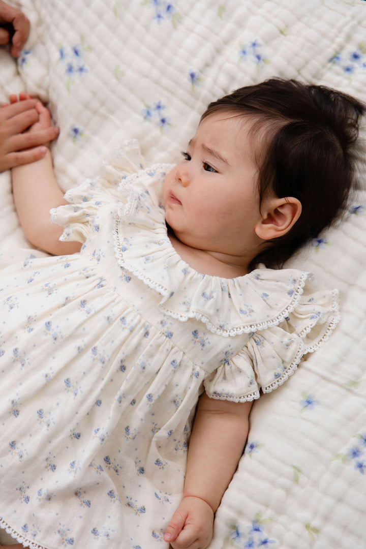 Baby in a white floral dress lying on a quilted surface