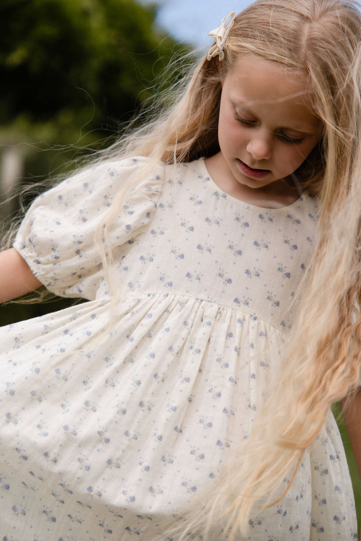 Young girl in a white floral dress standing outdoors with greenery in the background