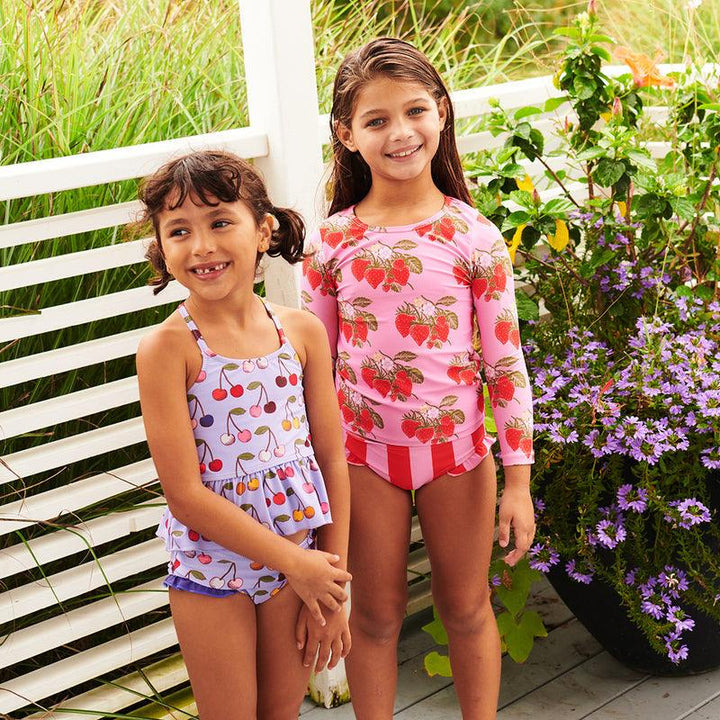 two little girls wearing swimsuits, one in a two piece with cherries on it and the other with the strawberries on it. the strawberry suit has a rashguard. 