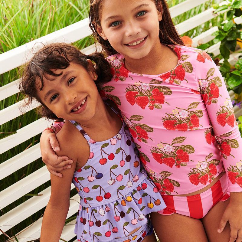 two little girls wearing swimsuits, one in a cherry two piece swim suit and the other in a strawberry two piece. the top is a rashguard. 
