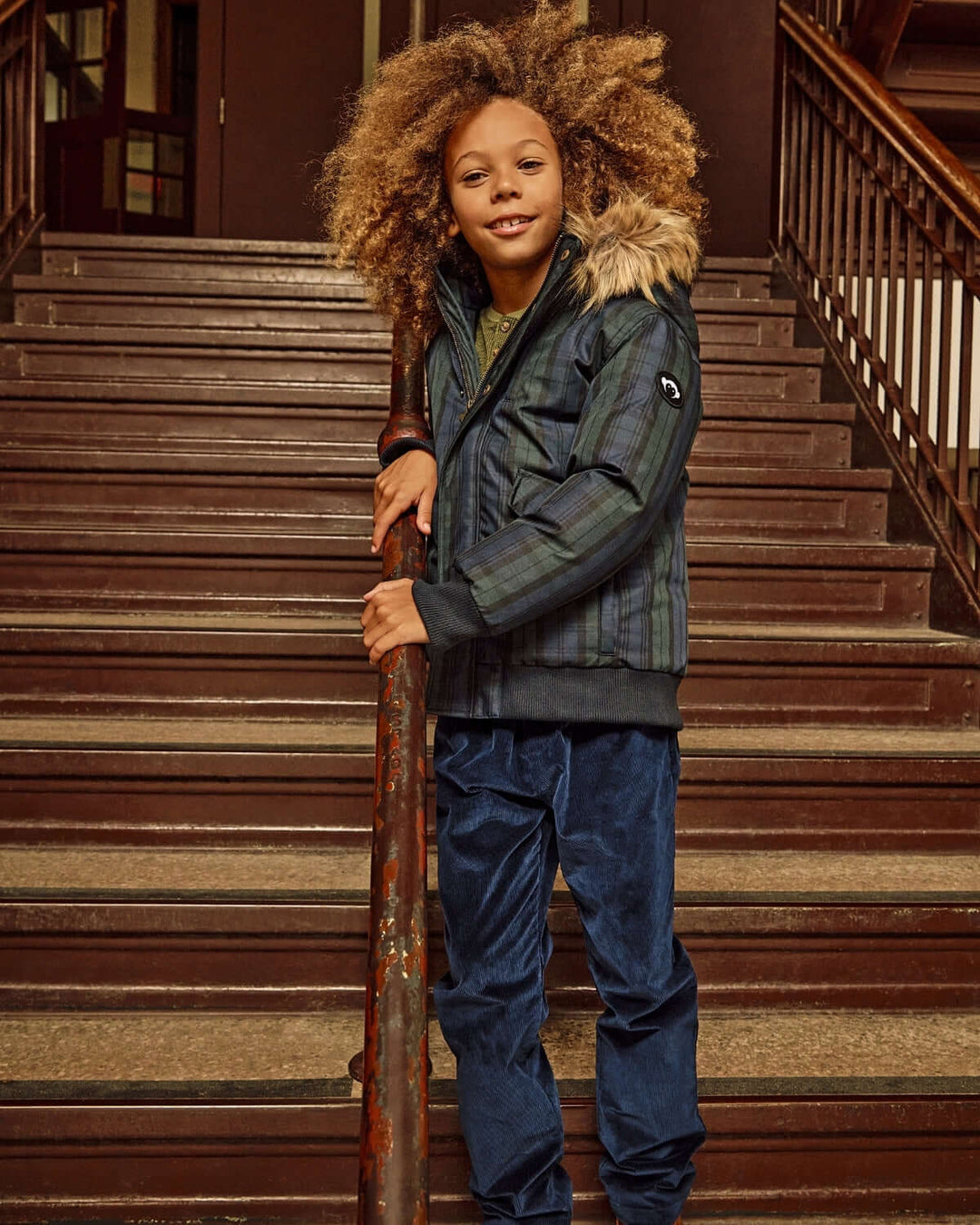 Child standing on a staircase wearing a green jacket with fur-lined hood and blue pants.
