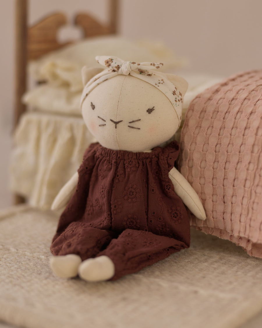 A plush cat doll wearing a dark brown eyelet dress and a cream bow headband, posed on a textured beige blanket next to a pink pillow.