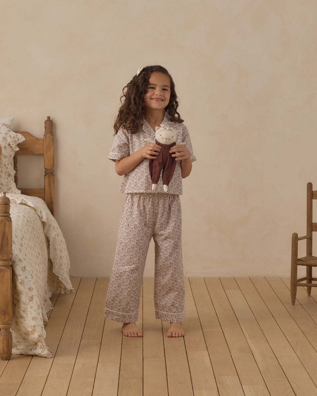 A young girl stands barefoot in a softly lit bedroom wearing a matching short-sleeve pajama top and wide-leg pants set with a small floral print. She is holding a plush doll, with a wooden bed and chair visible in the room.