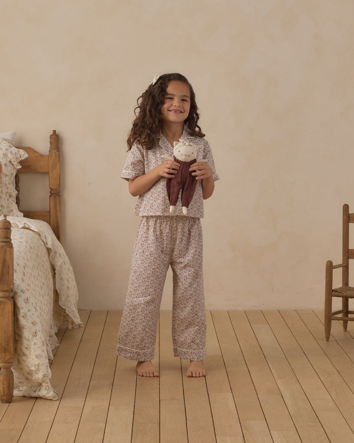 A young girl stands barefoot in a softly lit bedroom wearing a matching short-sleeve pajama top and wide-leg pants set with a small floral print. She is holding a plush doll, with a wooden bed and chair visible in the room.
