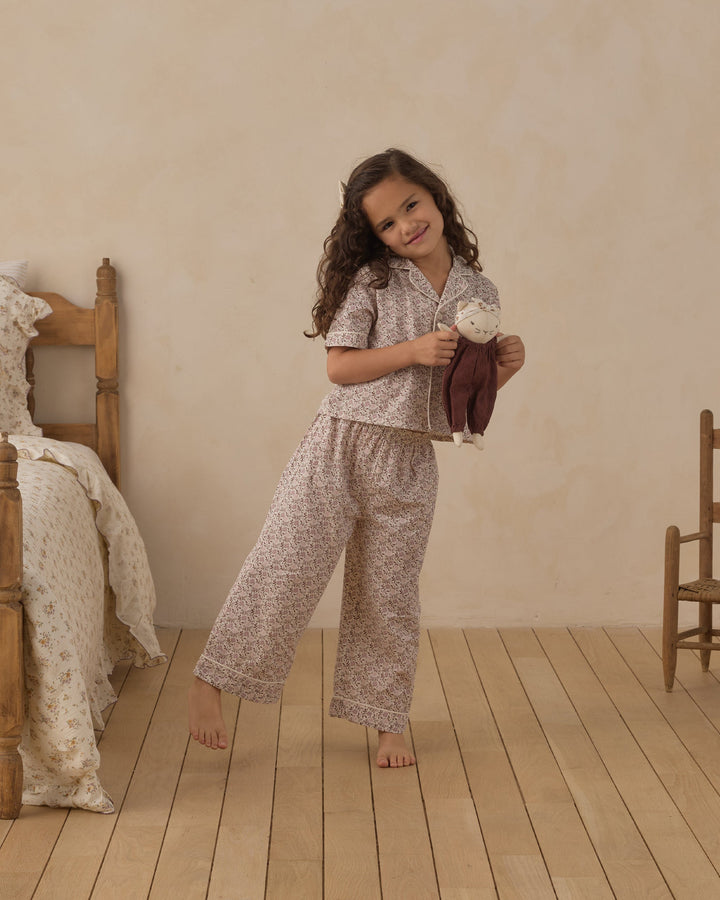 A young girl stands barefoot in a bedroom setting, wearing a matching short-sleeve button-up pajama top and wide-leg pants set in a light beige and white floral pattern. She is holding a brown plush toy.