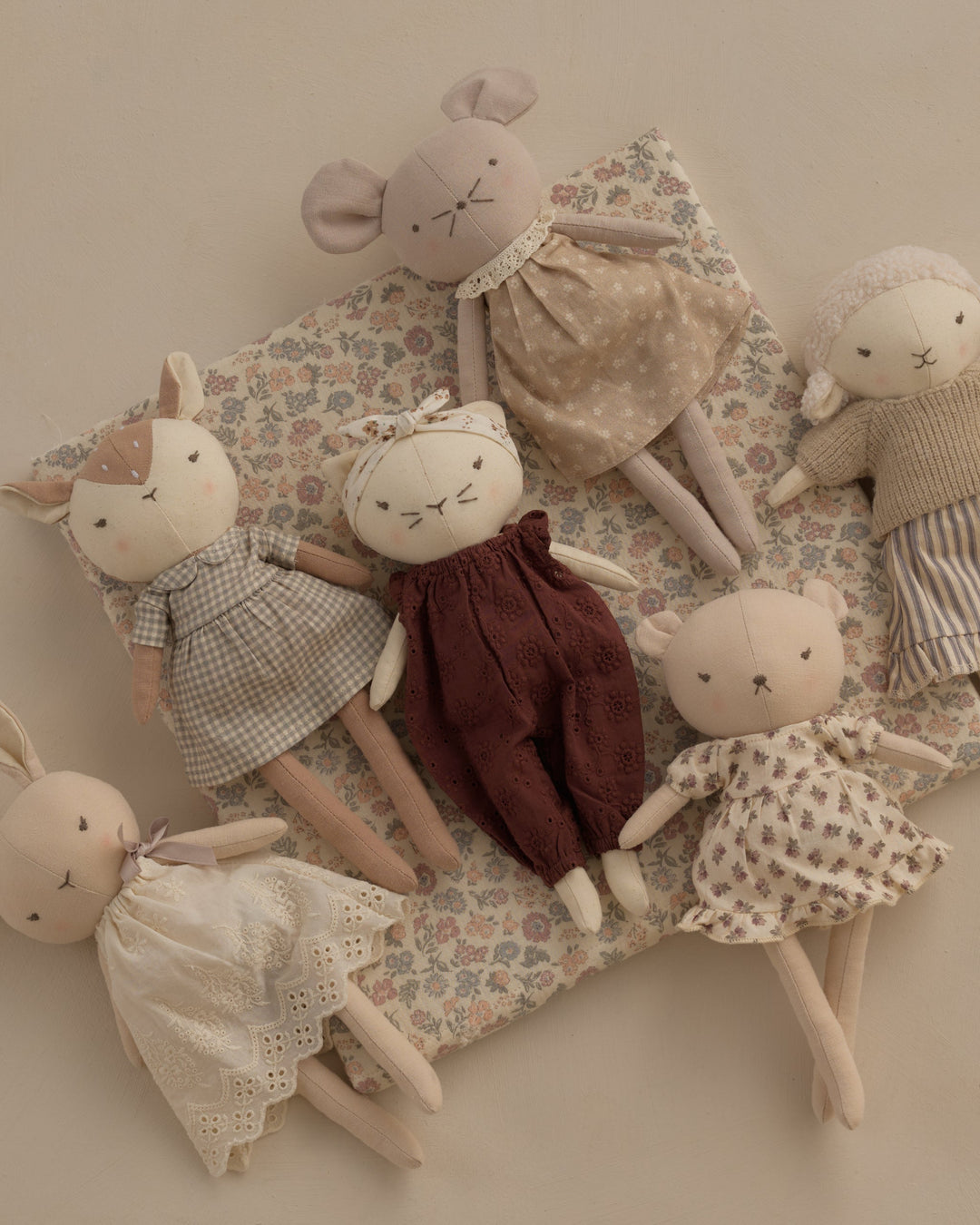 A set of five handmade fabric animal dolls, including a mouse, rabbit, sheep, and piglets, arranged on a floral patterned cushion against a warm beige background.