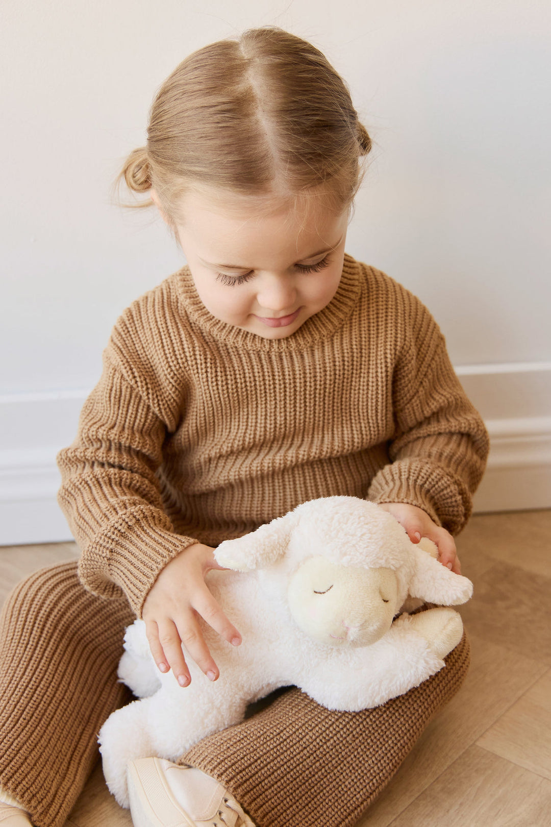 Child in a brown sweater holding a white plush lamb toy on a wooden floor.