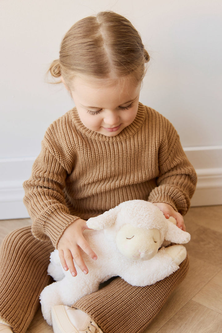 Child in a brown sweater holding a white plush lamb toy on a wooden floor.