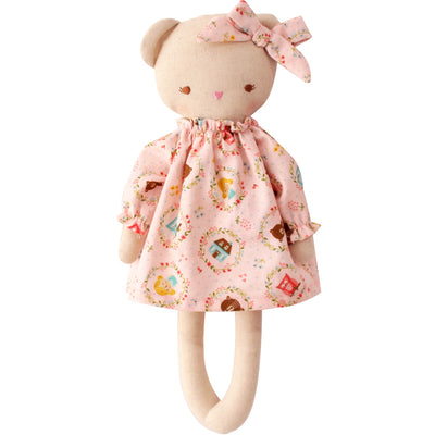 beige linen teddy bear dressed in a pink dress with little cottages and dolls printed on it. 