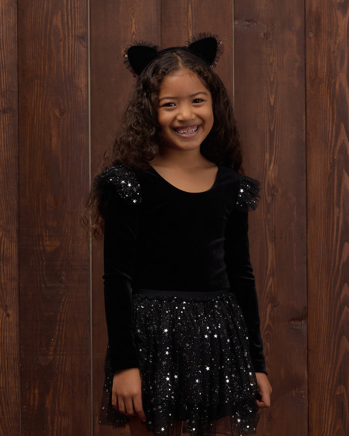 A young girl smiling in a black long-sleeve velvet top with sequined shoulders and a black sequined skirt, wearing black cat ears, standing against a dark brown wooden plank background.