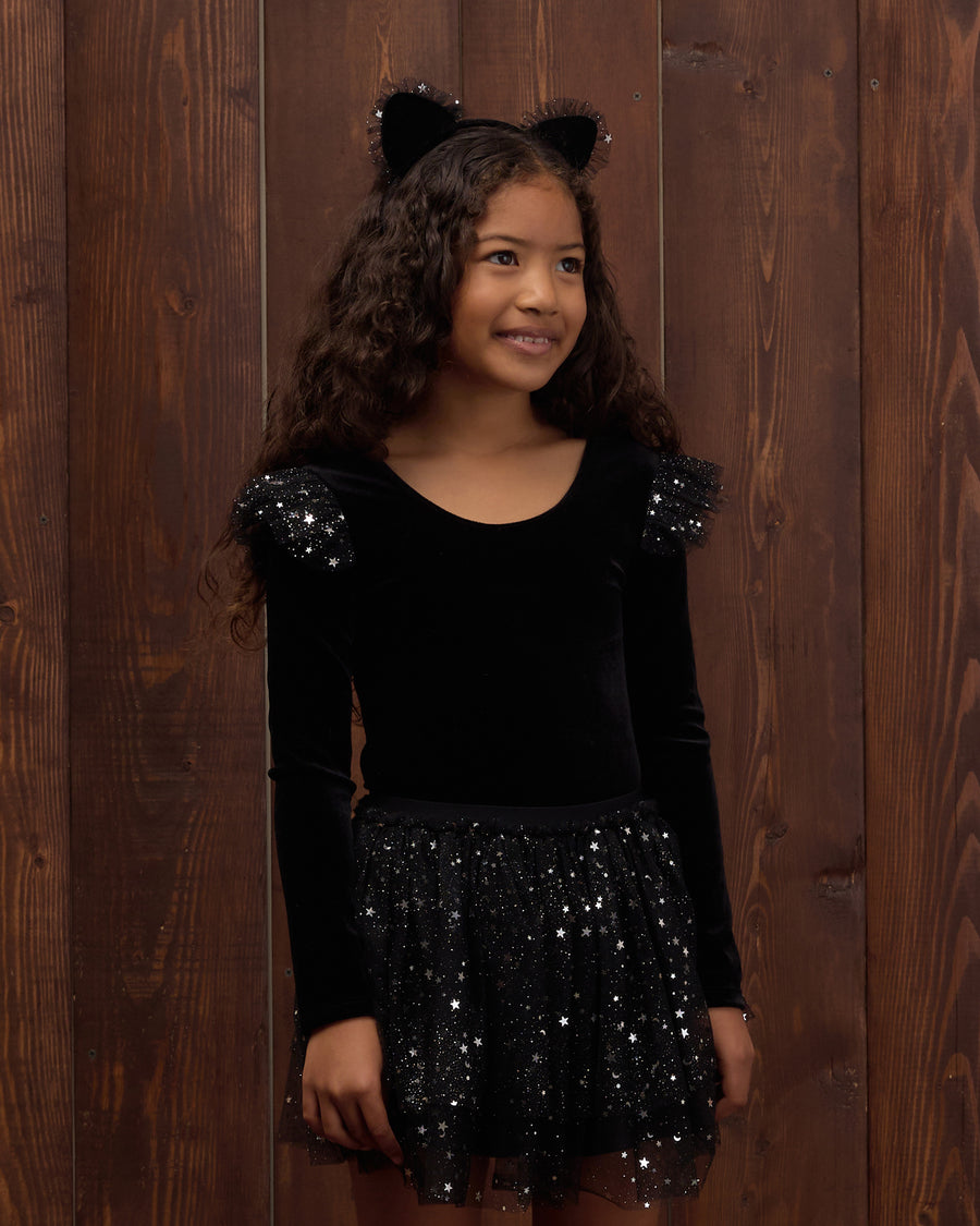 A young girl with long curly brown hair wearing a black velvet long-sleeve top with sequined shoulder details and a black sequined skirt, along with a black cat-ear headband, standing against a dark wood plank wall.