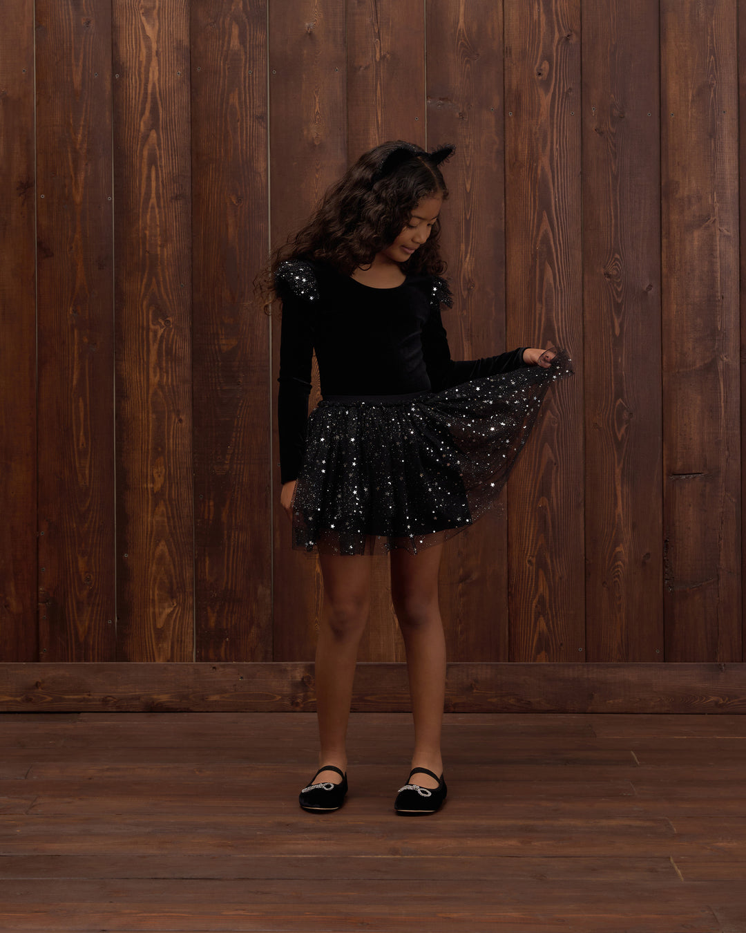 A young girl with curly brown hair stands in front of a dark wood-paneled wall, wearing a black long-sleeve top with sequined shoulders and a black sequined tutu skirt, holding out the skirt to show its sparkle. She is wearing black Mary Jane shoes.