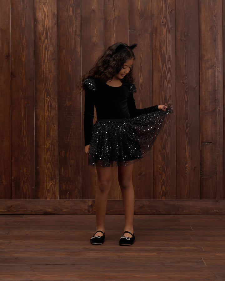 A young girl with curly brown hair stands in front of a dark wood-paneled wall, wearing a black long-sleeve top with sequined shoulders and a black sequined tutu skirt, holding out the skirt to show its sparkle. She is wearing black Mary Jane shoes.