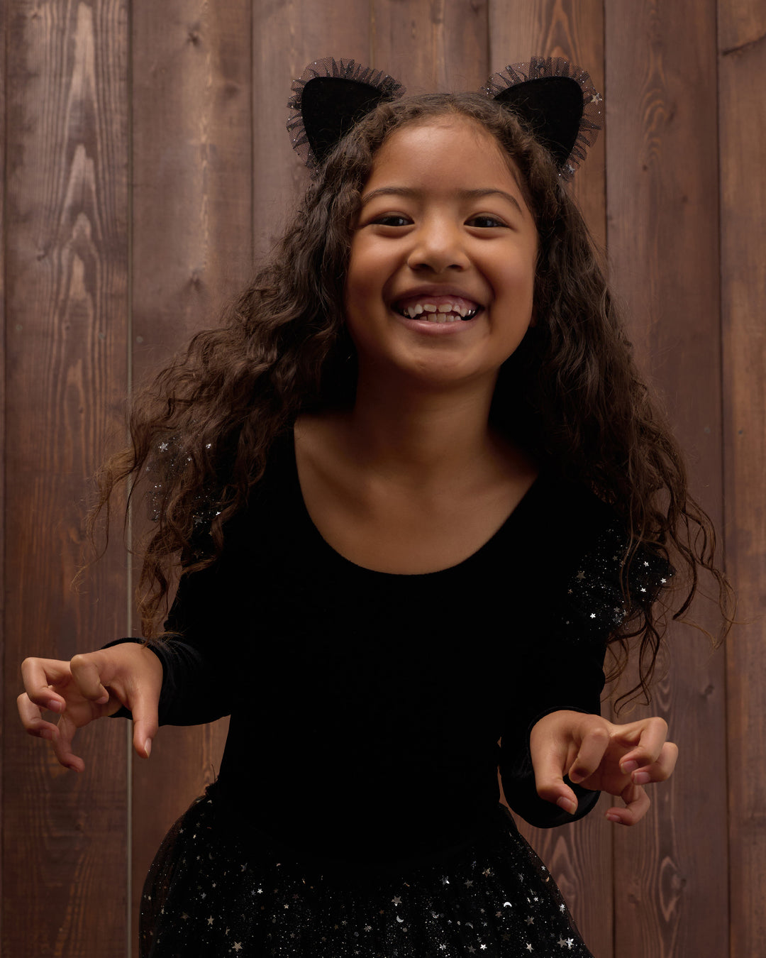 A young girl with long curly brown hair wearing a black cat costume top with attached cat ears and a sparkly black skirt, posing in front of a dark wood plank background.